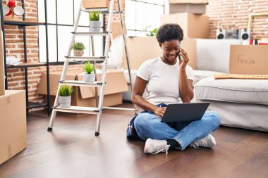 Afro-Amerikalı kadın dizüstü bilgisayar kullanıyor ve akıllı telefonla konuşuyor. Yeni evinde yerde oturuyor.