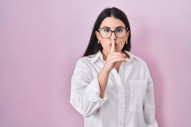 Genç esmer kadın pembe arka planda dikiliyor ve parmakları dudaklarında sessiz olmak istiyor. sessizlik ve gizli konsept. 