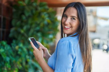 Genç, güzel, İspanyol bir kadın sokakta akıllı telefon kullanarak gülümsüyor.