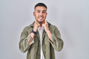 Soyutlanmış arka planda duran genç İspanyol adam ağzı açık gülümsüyor, parmakları işaret ediyor ve neşeli bir gülümseme sergiliyor. 