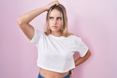 Young blonde woman standing over pink background confuse and wondering about question. uncertain with doubt, thinking with hand on head. pensive concept. 