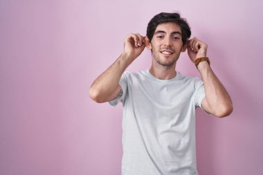 Young hispanic man standing over pink background smiling pulling ears with fingers, funny gesture. audition problem 