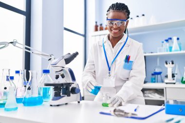 African american woman scientist smiling confident pouring liquid on test tube at laboratory