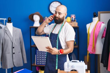 Young bald man tailor talking on smartphone looking clothing design at clothing factory