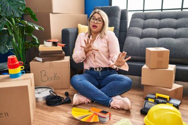 Young hispanic woman moving to a new home sitting on the floor afraid and terrified with fear expression stop gesture with hands, shouting in shock. panic concept. 