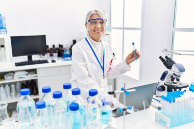 Orta yaşlı, gri saçlı, bilim adamı üniforması giyen bir kadın laboratuarda dizüstü bilgisayar kullanarak test tüpü tutuyor.