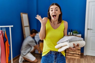 Young brunette woman holding folded laundry celebrating victory with happy smile and winner expression with raised hands 