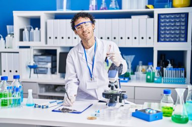Young hispanic man scientist writing on document holding blood test tube at laboratory