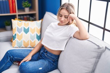 Sarışın genç kadın ellerini başının üstüne koyup evdeki kanepede oturuyor.