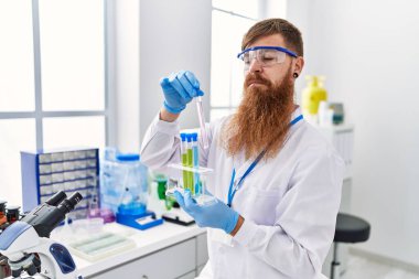 Laboratuvarda test tüpleri tutan bilim adamı üniforması giyen genç kızıl saçlı adam.