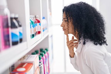 Young hispanic woman customer looking bottle on shelving at pharmacy