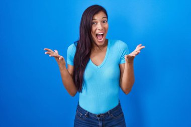 Young hispanic woman standing over blue background celebrating crazy and amazed for success with arms raised and open eyes screaming excited. winner concept 