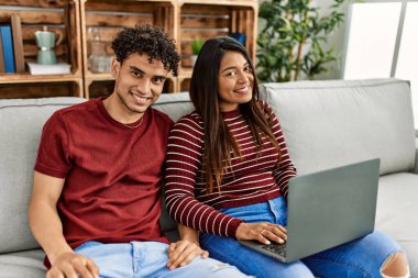 Evdeki kanepede oturmuş dizüstü bilgisayar kullanarak mutlu bir şekilde gülümseyen genç Latin çift..