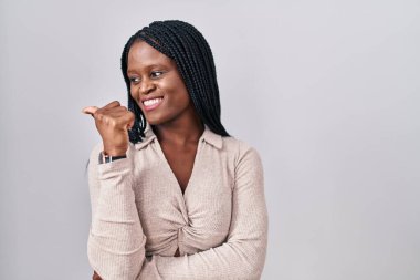 African woman with braids standing over white background smiling with happy face looking and pointing to the side with thumb up. 