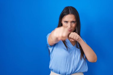 Young brunette woman standing over blue background punching fist to fight, aggressive and angry attack, threat and violence 