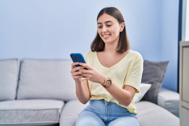 Evdeki kanepede oturan, akıllı telefon kullanan genç İspanyol kadın.