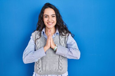 Genç esmer kadın mavi arka planda duruyor. Elleri birarada dua ediyor. Affedilmek istiyor.. 