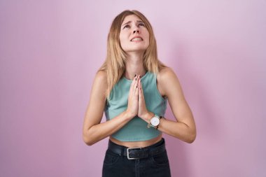 Sarışın, beyaz bir kadın pembe arka planda duruyor. Elleriyle yalvarıyor ve dua ediyor. Yüzünde umut ifadesi var. Yalvarıyor. 