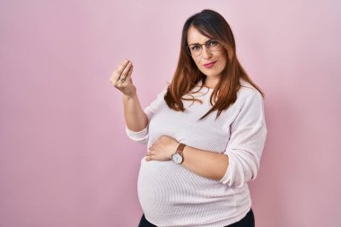 Pregnant woman standing over pink background doing italian gesture with hand and fingers confident expression 