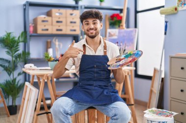 Arab man with beard painter sitting at art studio holding palette pointing finger to one self smiling happy and proud 