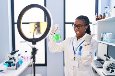 African woman with braids working at scientist laboratory doing tutorial with smartphone pointing thumb up to the side smiling happy with open mouth 
