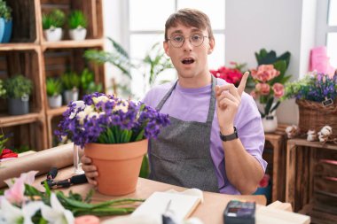 Caucasian blond man working at florist shop surprised pointing with finger to the side, open mouth amazed expression. 