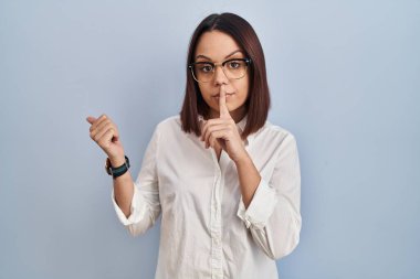 Young hispanic woman standing over white background asking to be quiet with finger on lips pointing with hand to the side. silence and secret concept. 