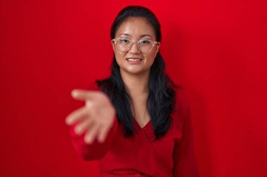 Asian young woman standing over red background smiling cheerful offering palm hand giving assistance and acceptance. 