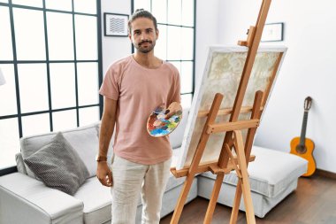 Young hispanic man with beard painting on canvas at home relaxed with serious expression on face. simple and natural looking at the camera. 