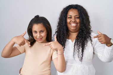 Anne ve küçük kızı beyaz arka planda duruyor. Yüzünde bir gülümsemeyle, kendini gururlu ve mutlu olarak gösteriyor.. 