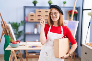 Young redhead woman at art studio holding art case gesturing finger crossed smiling with hope and eyes closed. luck and superstitious concept. 