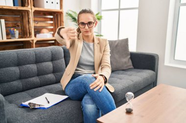 Young woman working at consultation office looking unhappy and angry showing rejection and negative with thumbs down gesture. bad expression. 