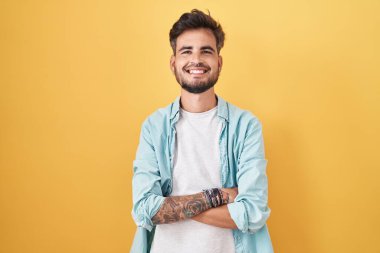 Sarı arka planda dikilen dövmeli genç İspanyol adam kollarını kavuşturup kameraya bakarken gülümsüyor. pozitif kişi. 