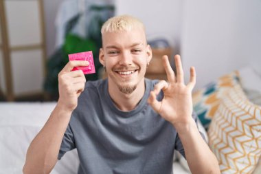 Kafkasyalı genç bir adam elinde prezervatifle yatakta oturmuş parmaklarıyla işaret veriyor, gülümsüyor ve mükemmel bir sembol oluşturuyor. 