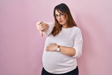 Pregnant woman standing over pink background looking unhappy and angry showing rejection and negative with thumbs down gesture. bad expression. 