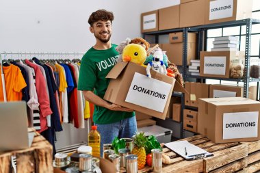 Young arab man wearing volunteer uniform holding donations box with toys at charity center