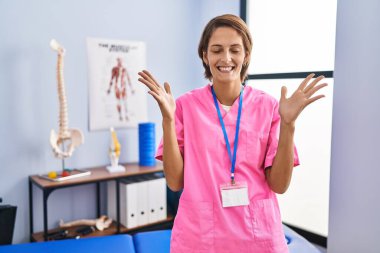 Brunette woman working at rehabilitation clinic celebrating mad and crazy for success with arms raised and closed eyes screaming excited. winner concept 