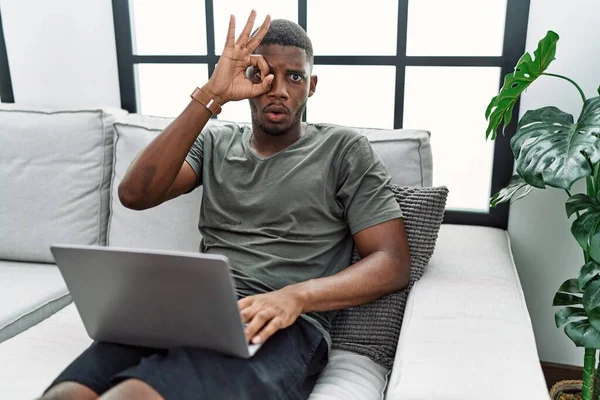 Young african american man using laptop at home sitting on the sofa doing ok gesture shocked with surprised face, eye looking through fingers. unbelieving expression.
