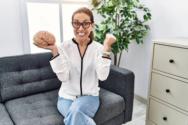 Young hispanic woman holding brain working at psychology clinic screaming proud, celebrating victory and success very excited with raised arms
