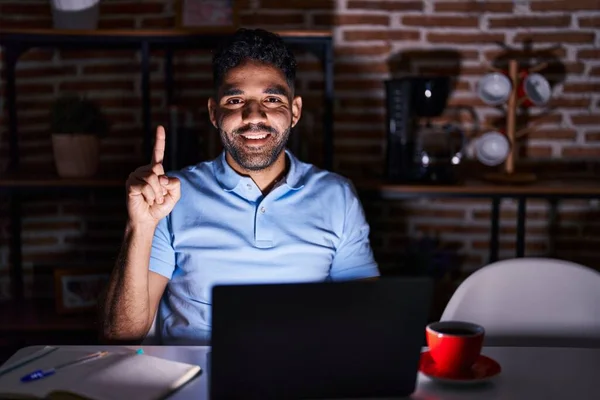 Hispanic man with beard using laptop at night showing and pointing up with finger number one while smiling confident and happy.