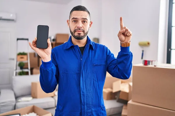 Hispanic man working on moving service showing smartphone screen smiling with an idea or question pointing finger with happy face, number one