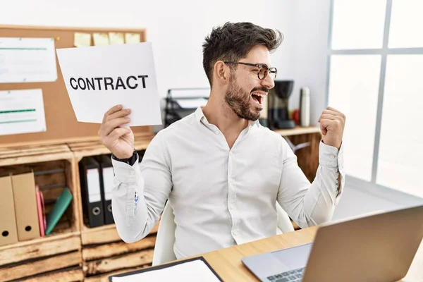 Young man with beard holding contract paper at the office pointing thumb up to the side smiling happy with open mouth