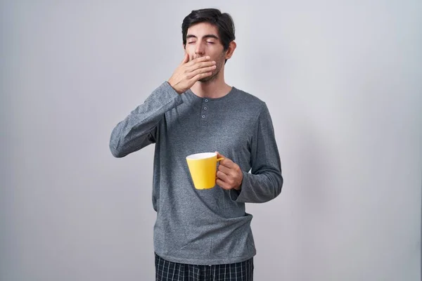 Young hispanic man wearing pajama drinking a cup of coffee bored yawning tired covering mouth with hand. restless and sleepiness.