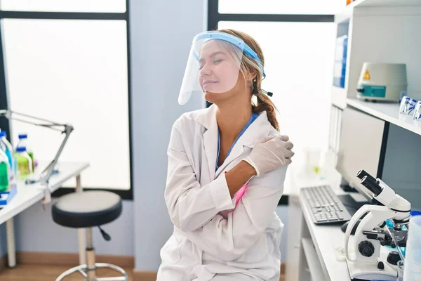 Young blonde woman working at scientist laboratory wearing face mask hugging oneself happy and positive, smiling confident. self love and self care