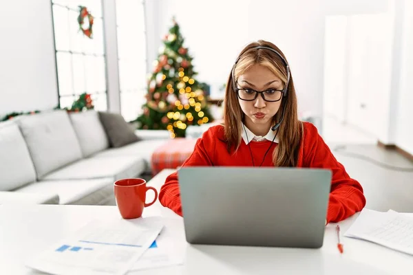 年轻的高加索女孩坐在桌子上 用笔记本电脑在圣诞树旁工作 脸上挂着滑稽的表情 气喘吁吁的嘴 疯狂的表情 — 图库照片