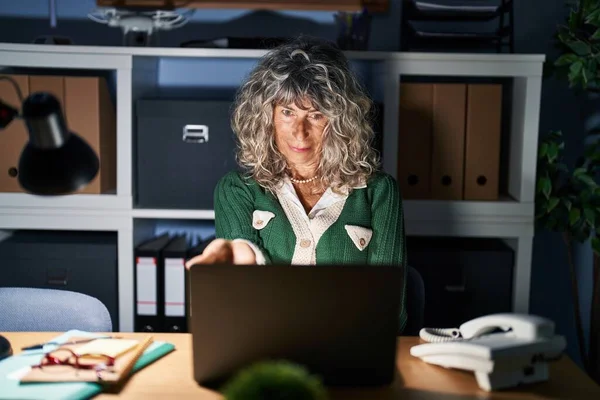 Middle age woman working at night using computer laptop smiling cheerful offering palm hand giving assistance and acceptance.