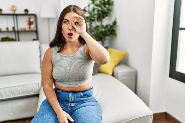 Young brunette woman sitting on the sofa at home doing ok gesture shocked with surprised face, eye looking through fingers. unbelieving expression.