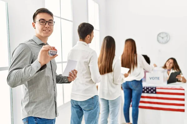Group of young american voter people putting vote in ballot. Man smiling happy and holding i voted badge at electoral center.