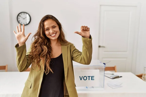 Beautiful hispanic woman standing at political campaign room showing and pointing up with fingers number six while smiling confident and happy.