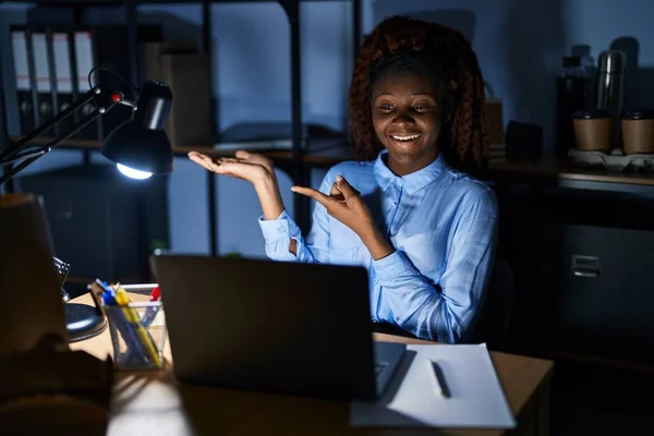 African woman working at the office at night amazed and smiling to the camera while presenting with hand and pointing with finger.